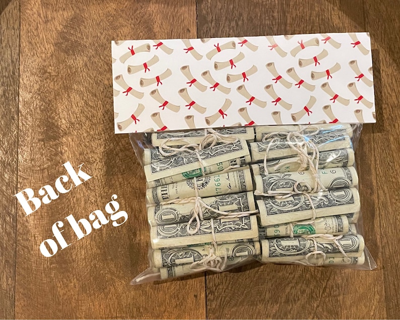 May include: A clear plastic bag filled with rolled up dollar bills tied with twine. The bag is sitting on a wooden surface. The bag is decorated with a white and red patterned paper with a design of red and white diplomas.