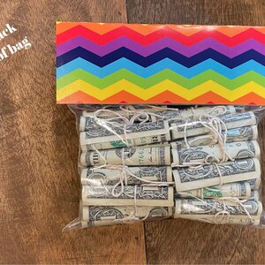 May include: A clear plastic bag filled with rolled up US dollar bills tied with string. The bag is sitting on a wooden surface. The bag is decorated with a rainbow chevron pattern.