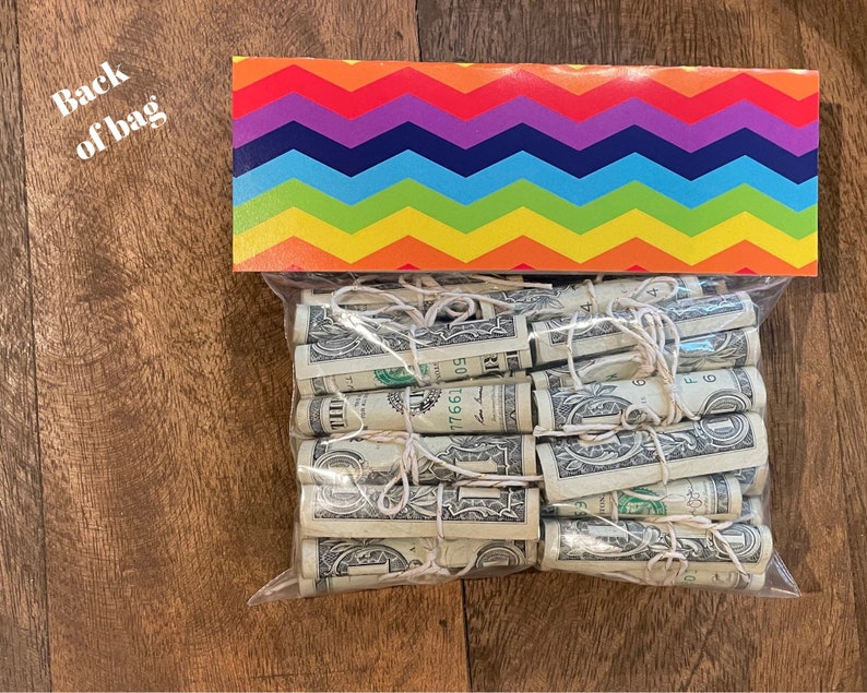 May include: A clear plastic bag filled with rolled up US dollar bills tied with string. The bag is sitting on a wooden surface. The bag is decorated with a rainbow chevron pattern.