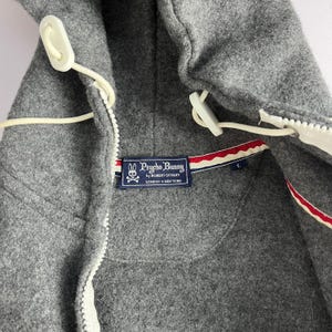 Puede incluir: Primer plano de una chaqueta con capucha Psycho Bunny gris. La chaqueta presenta una cremallera blanca, cierres de palanca y una etiqueta azul marino con el logotipo y el texto de la marca. La etiqueta tiene detalles a rayas rojas y blancas.