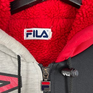 Puede incluir: Primer plano de una sudadera con cremallera Fila. La sudadera presenta un dise&ntilde;o de bloques de color gris, negro y rojo con forro polar rojo. El logotipo de Fila es visible en el tirador de la cremallera y en la etiqueta interior. La sudadera tiene un cord&oacute;n y una cremallera completa.