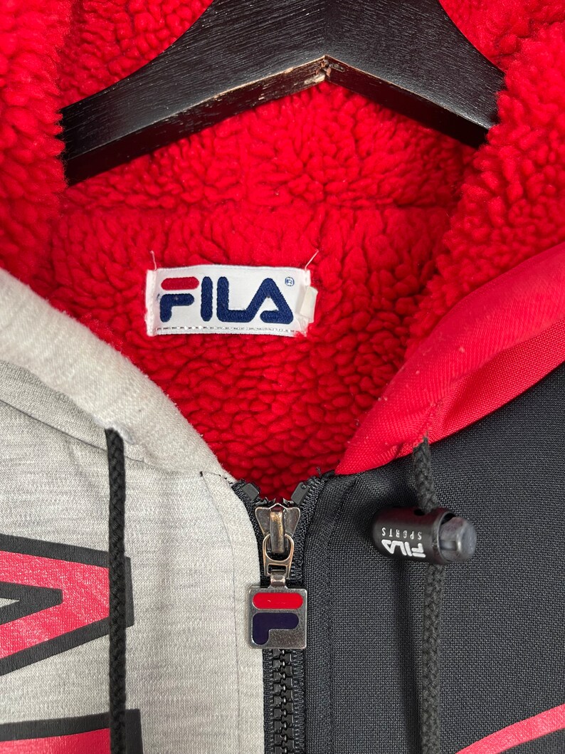 Puede incluir: Primer plano de una sudadera con cremallera Fila. La sudadera presenta un dise&ntilde;o de bloques de color gris, negro y rojo con forro polar rojo. El logotipo de Fila es visible en el tirador de la cremallera y en la etiqueta interior. La sudadera tiene un cord&oacute;n y una cremallera completa.