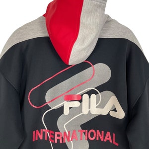 Puede incluir: Sudadera con capucha negra con capucha de bloques de color rojo y gris. La parte trasera presenta un gran gr&aacute;fico con el nombre de la marca "FILA" en blanco y rojo, junto con la palabra "INTERNATIONAL" en rojo.
