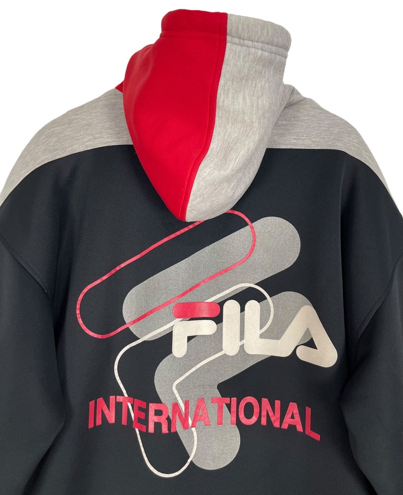 Puede incluir: Sudadera con capucha negra con capucha de bloques de color rojo y gris. La parte trasera presenta un gran gr&aacute;fico con el nombre de la marca "FILA" en blanco y rojo, junto con la palabra "INTERNATIONAL" en rojo.