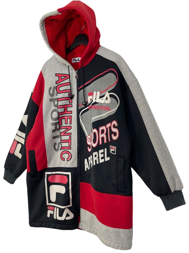Puede incluir: Una sudadera con capucha con cremallera Fila en rojo, negro y gris. La chaqueta presenta el logotipo de Fila y las palabras "Authentic Sports Apparel". La capucha es roja con un forro suave. Tiene cremallera frontal y dos bolsillos.