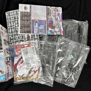 Puede incluir: Un conjunto de piezas de un kit de modelo Gundam sin montar, que incluye marcos de pl&aacute;stico, manuales de instrucciones y calcoman&iacute;as. El embalaje presenta el texto "RX-78F00 GUNDAM & G-DOCK" y el logotipo "BANDAI".