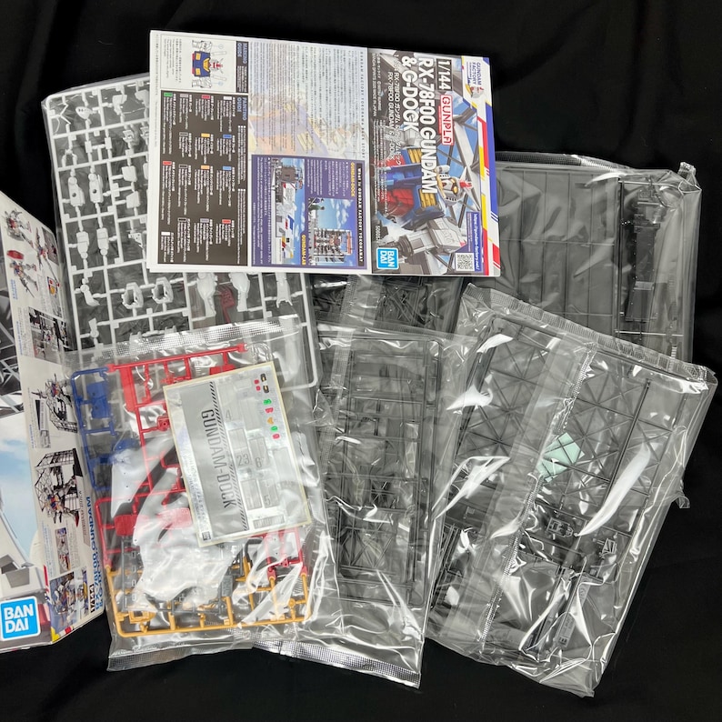 Puede incluir: Un conjunto de piezas de un kit de modelo Gundam sin montar, que incluye marcos de pl&aacute;stico, manuales de instrucciones y calcoman&iacute;as. El embalaje presenta el texto "RX-78F00 GUNDAM & G-DOCK" y el logotipo "BANDAI".