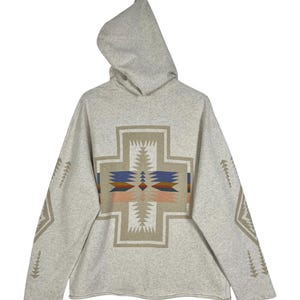 May include: A light grey hooded sweatshirt with a large cross-shaped design on the front. The cross features geometric patterns in beige, blue, and orange. The sleeves have geometric patterns in beige. The sweatshirt has a relaxed fit.