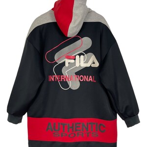 Puede incluir: Chaqueta con capucha Fila negra, roja y gris. La chaqueta presenta el logotipo de Fila y las palabras "INTERNATIONAL" y "AUTHENTIC SPORTS" en rojo. La capucha es roja y gris.