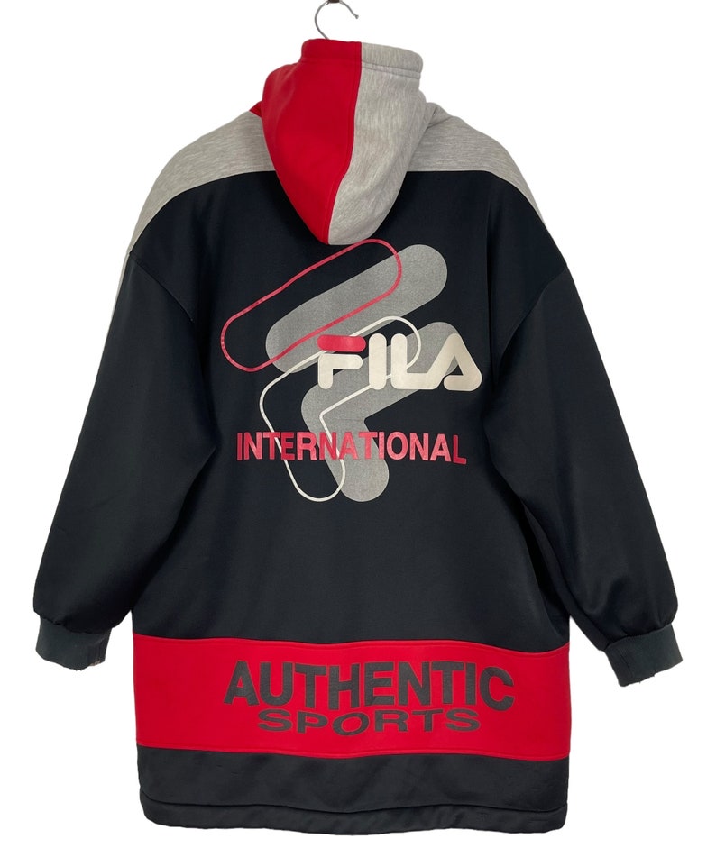 Puede incluir: Chaqueta con capucha Fila negra, roja y gris. La chaqueta presenta el logotipo de Fila y las palabras "INTERNATIONAL" y "AUTHENTIC SPORTS" en rojo. La capucha es roja y gris.