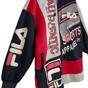 Puede incluir: Una sudadera con capucha con cremallera y bloques de color con el logotipo de FILA y "AUTHENTIC" impreso verticalmente. La chaqueta presenta paneles negros, rojos y grises con forro interior rojo. Tambi&eacute;n est&aacute;n impresas las palabras "SPORTS APPAREL".