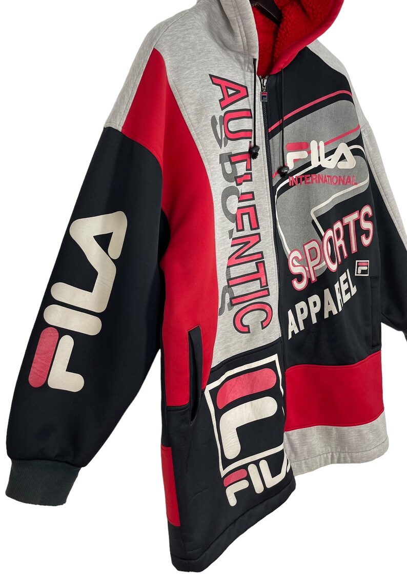 Puede incluir: Una sudadera con capucha con cremallera y bloques de color con el logotipo de FILA y "AUTHENTIC" impreso verticalmente. La chaqueta presenta paneles negros, rojos y grises con forro interior rojo. Tambi&eacute;n est&aacute;n impresas las palabras "SPORTS APPAREL".