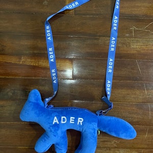 May include: A blue plush fox-shaped bag with a blue strap that says "ADER" repeatedly. The bag has a zipper and the word "ADER" is embroidered on the front.