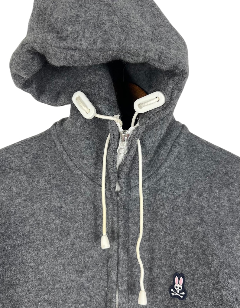 Puede incluir: Una sudadera con capucha gris con cierre de cremallera completo y cordones blancos. La capucha es grande y la sudadera tiene un peque&ntilde;o logotipo bordado de un conejito y una calavera en el pecho.