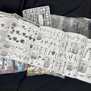 Puede incluir: Un manual de instrucciones abierto para un kit de modelo, con diagramas detallados y n&uacute;meros de piezas. Se ven piezas de modelo de pl&aacute;stico, junto con bolsas de pl&aacute;stico transparente y una caja con el logotipo "BANDAI".