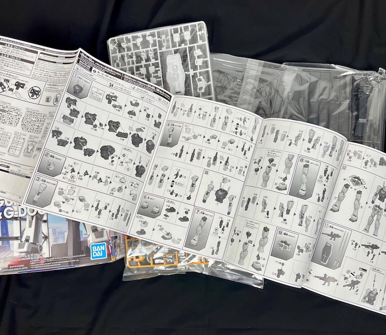 Puede incluir: Un manual de instrucciones abierto para un kit de modelo, con diagramas detallados y n&uacute;meros de piezas. Se ven piezas de modelo de pl&aacute;stico, junto con bolsas de pl&aacute;stico transparente y una caja con el logotipo "BANDAI".