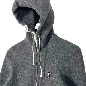 Puede incluir: Una sudadera con capucha gris con cierre de cremallera completo y cordones blancos. La sudadera presenta un peque&ntilde;o logotipo bordado de un conejo con una calavera y huesos cruzados en el pecho. La capucha est&aacute; forrada con tela blanca.