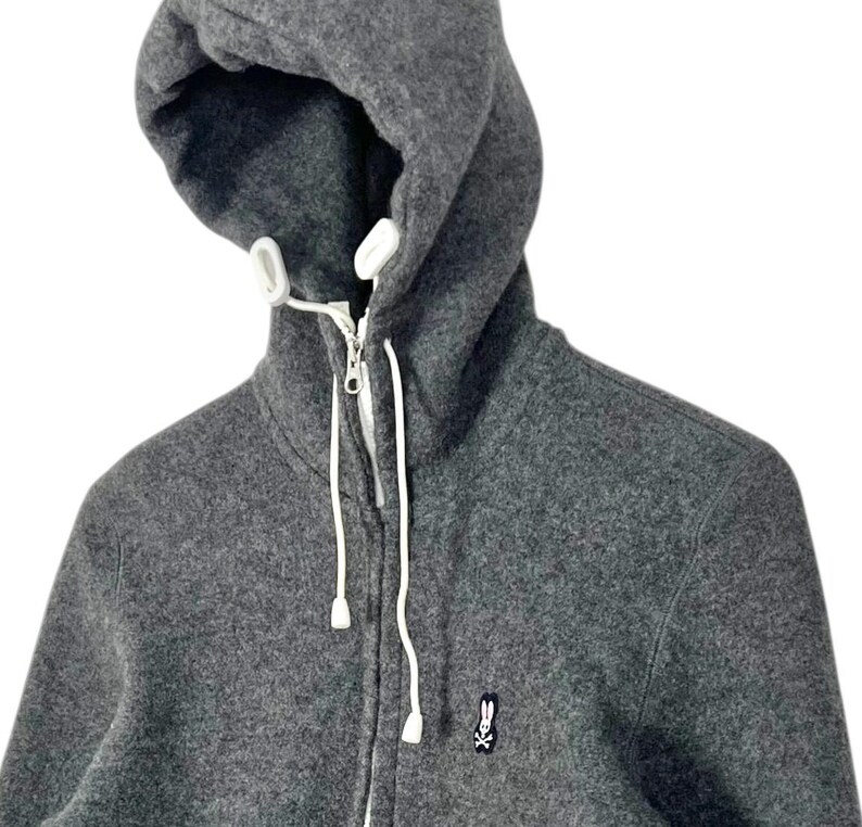 Puede incluir: Una sudadera con capucha gris con cierre de cremallera completo y cordones blancos. La sudadera presenta un peque&ntilde;o logotipo bordado de un conejo con una calavera y huesos cruzados en el pecho. La capucha est&aacute; forrada con tela blanca.