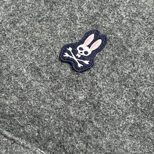 Puede incluir: Primer plano de una tela de punto gris. Se ve un peque&ntilde;o parche bordado, con una calavera blanca y huesos cruzados con una cabeza de conejo rosa, contorneada en blanco y azul marino.