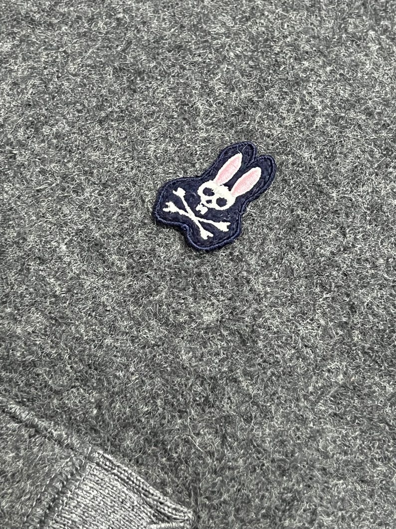 Puede incluir: Primer plano de una tela de punto gris. Se ve un peque&ntilde;o parche bordado, con una calavera blanca y huesos cruzados con una cabeza de conejo rosa, contorneada en blanco y azul marino.