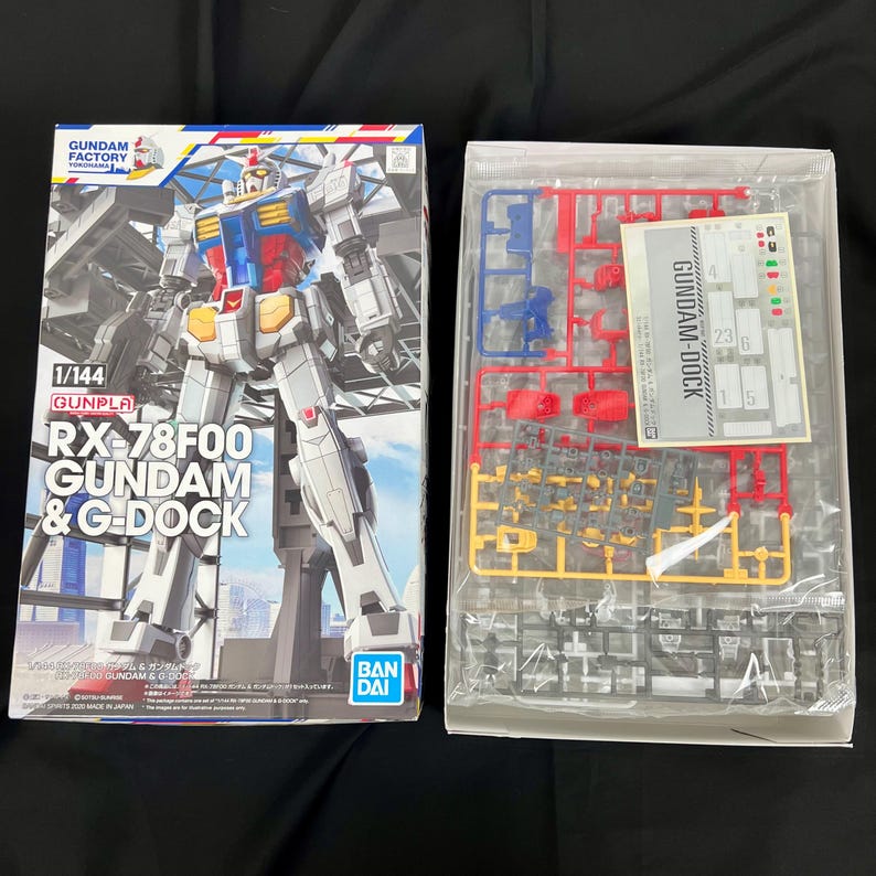 Puede incluir: Un kit de modelo RX-78F00 Gundam & G-Dock en caja. La caja presenta una ilustraci&oacute;n detallada del robot Gundam en blanco, azul, rojo y amarillo. El kit incluye piezas de pl&aacute;stico de varios colores y una hoja de calcoman&iacute;as.