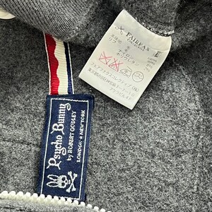 Puede incluir: Primer plano de una etiqueta de sudadera con capucha Psycho Bunny gris. La etiqueta presenta el nombre de la marca, un logotipo de calavera de conejo y el texto "LONDON * NEW YORK". Tambi&eacute;n es visible una etiqueta blanca con texto japon&eacute;s.