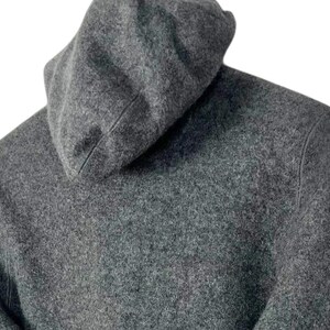 Puede incluir: Primer plano de una sudadera con capucha gris. La capucha est&aacute; levantada y la tela tiene un aspecto texturizado. La sudadera es de color gris liso y el fondo es blanco.