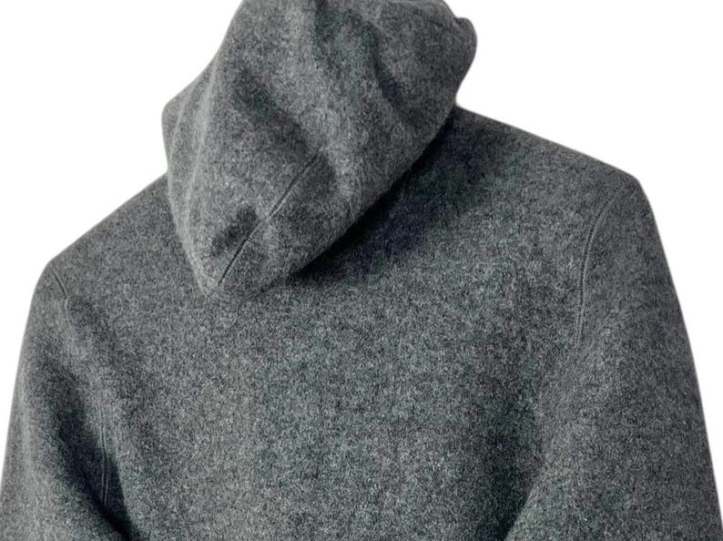 Puede incluir: Primer plano de una sudadera con capucha gris. La capucha est&aacute; levantada y la tela tiene un aspecto texturizado. La sudadera es de color gris liso y el fondo es blanco.