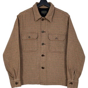 Può includere: Giacca a camicia a quadri marrone e rossa con due tasche sul petto e chiusure con bottoni. La camicia ha un colletto e polsini abbottonati. Il marchio "Pendleton" è visibile su un'etichetta vicino al colletto.