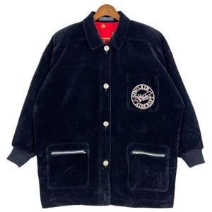 Może przedstawiać: Czarna sztruksowa kurtka z zapięciem na guziki, dwiema kieszeniami na zamek i naszywką na piersi z napisem "Sinclair Aircraft". Kurtka ma czerwoną podszewkę i czarne prążkowane mankiety.