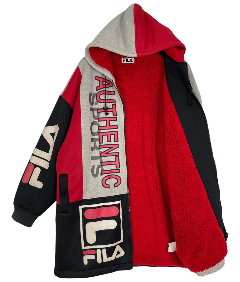 Puede incluir: Chaqueta larga con capucha Fila en negro, rojo y gris. La chaqueta presenta el logotipo de Fila y las palabras "AUTHENTIC SPORTS" en un dise&ntilde;o vertical. El interior est&aacute; forrado con forro polar rojo. La chaqueta tiene cierre de cremallera y capucha.