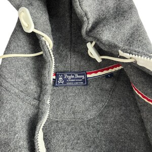 Puede incluir: Primer plano de una chaqueta con capucha gris con cremallera blanca y cierres de palanca. La chaqueta presenta una etiqueta Psycho Bunny con un logotipo y texto. El forro tiene un detalle a rayas rojas, blancas y azules.