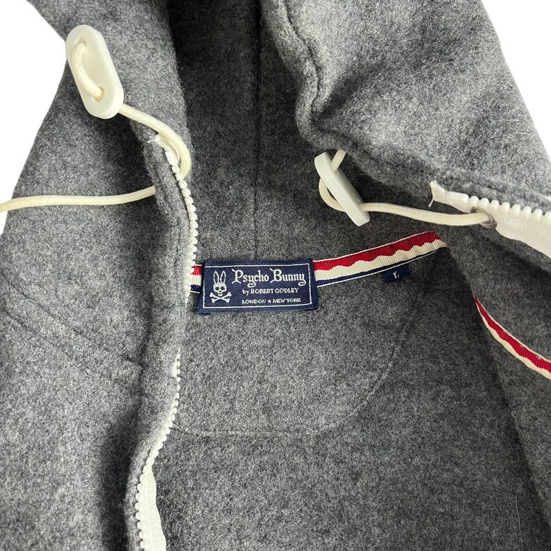 Puede incluir: Primer plano de una chaqueta con capucha gris con cremallera blanca y cierres de palanca. La chaqueta presenta una etiqueta Psycho Bunny con un logotipo y texto. El forro tiene un detalle a rayas rojas, blancas y azules.