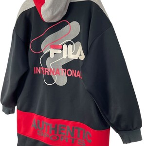 Puede incluir: Sudadera con capucha Fila negra y roja con capucha gris. La parte trasera presenta el logotipo de Fila y la palabra "INTERNATIONAL" en rojo. La parte inferior tiene una banda roja con las palabras "AUTHENTIC SPORTS".
