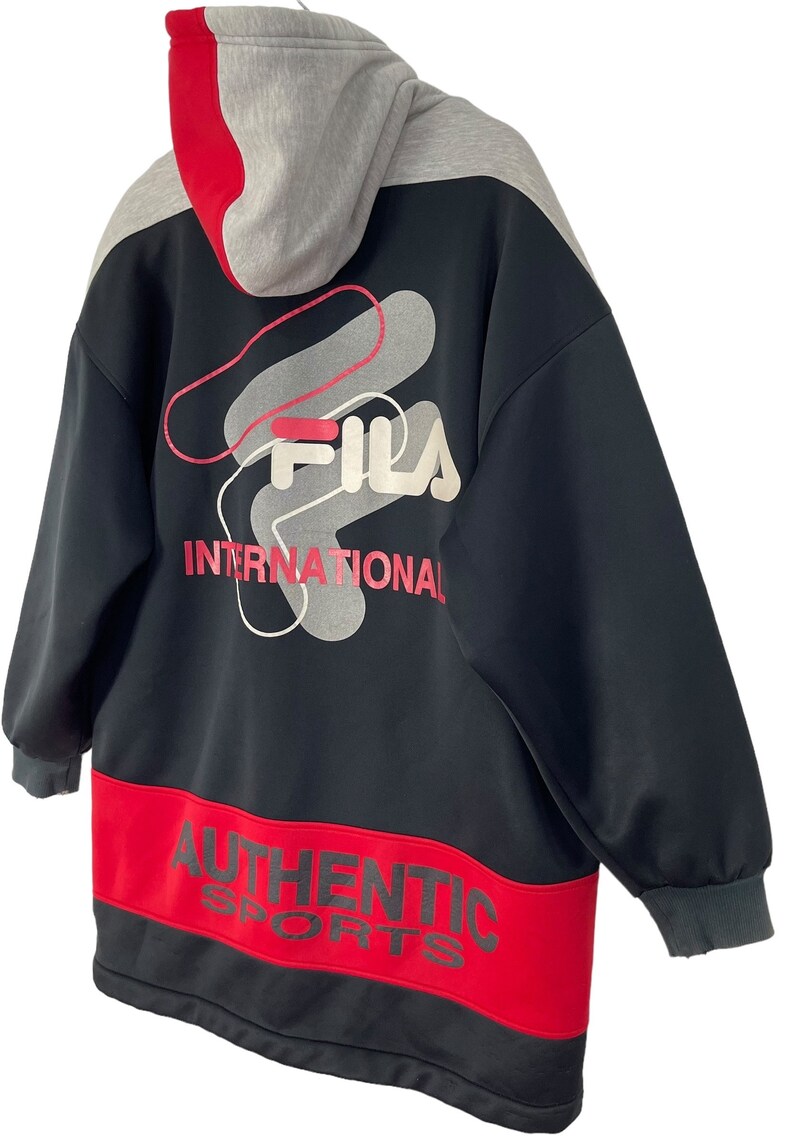 Puede incluir: Sudadera con capucha Fila negra y roja con capucha gris. La parte trasera presenta el logotipo de Fila y la palabra "INTERNATIONAL" en rojo. La parte inferior tiene una banda roja con las palabras "AUTHENTIC SPORTS".