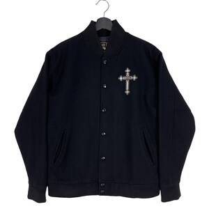 Puede incluir: Chaqueta de béisbol negra con una cruz plateada bordada en el pecho. La cruz tiene la palabra "Rock" escrita en ella.