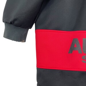 Puede incluir: Una sudadera gris oscuro con una banda roja en el pecho. La banda roja presenta la palabra "AU" en letras grandes de color gris oscuro. El pu&ntilde;o de la manga es de color gris claro.