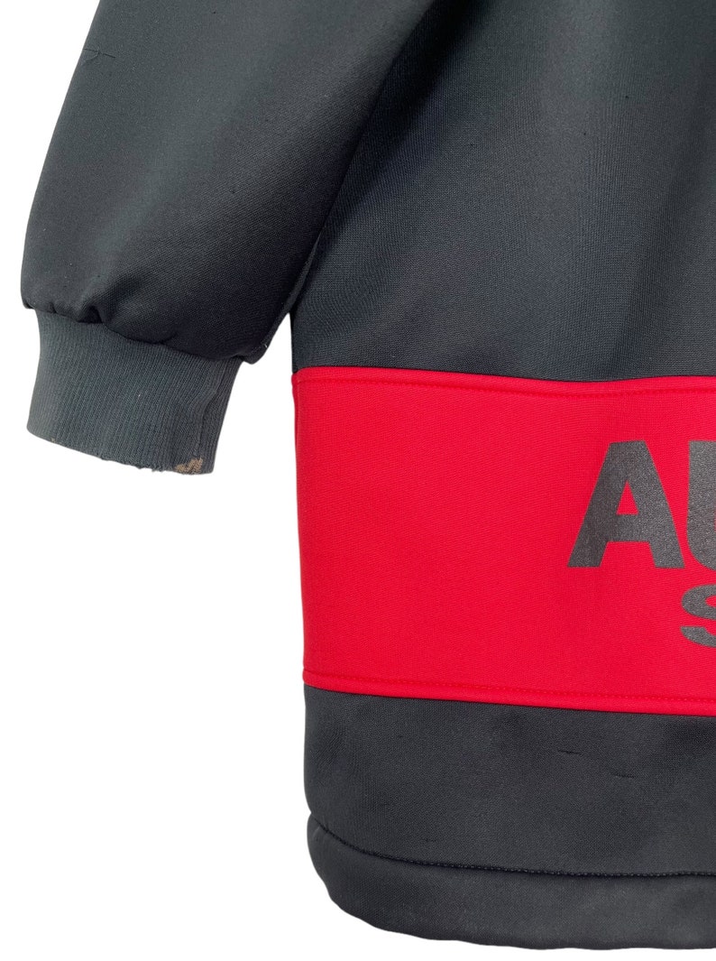 Puede incluir: Una sudadera gris oscuro con una banda roja en el pecho. La banda roja presenta la palabra "AU" en letras grandes de color gris oscuro. El pu&ntilde;o de la manga es de color gris claro.