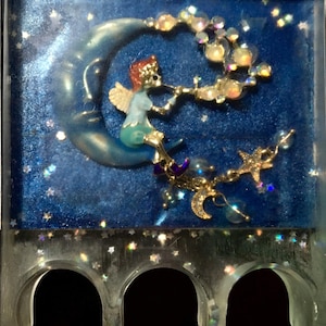 May include: A small, clear, resin figurine of a fairy sitting on a crescent moon. The fairy is wearing a blue dress and has a red crown. The moon is surrounded by a string of iridescent beads. The figurine is set against a blue background with silver glitter.