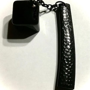 May include: A black keychain with a square charm and a textured, curved rectangular charm. The charms are connected by a small chain. The rectangular charm has a hammered metal appearance.