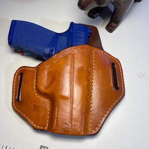 May include: A brown leather holster with a stitched design and a US flag embossed on the front. A blue handgun is partially visible in the holster.