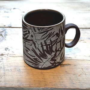 May include: A cylindrical ceramic mug featuring a dark brown interior and a handle. The exterior surface has a textured, light gray finish adorned with a repeating dark brown botanical pattern of elongated leaves and stems covering the entire body.