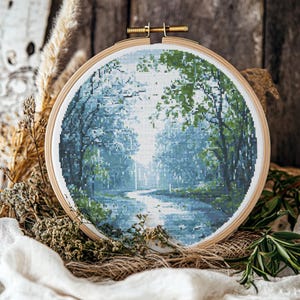 Puede incluir: Un bordado de punto de cruz que representa una escena de bosque con un río que lo atraviesa. El bordado está enmarcado en un aro de madera y presenta tonos de azul, verde y blanco.