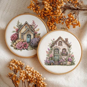 May include: Two cross-stitch embroidery hoops featuring detailed cottage scenes surrounded by flowers. One cottage is yellow with a blue door, the other is white with a green door. Both are set against a soft, neutral background.