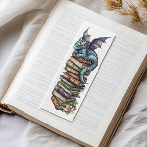 May include: A blue dragon bookmark with purple wings perched on a stack of colorful books. The bookmark is made of fabric and has a white background.