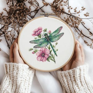 Puede incluir: Un bastidor de bordado de punto de cruz con una libélula verde y flores rosas. La libélula tiene alas detalladas y está rodeada de elementos florales. El bordado está en un bastidor de madera, sostenido por manos con un suéter color crema.