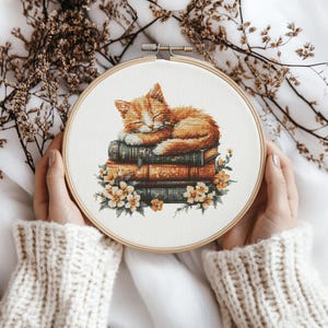 May include: A cross-stitch embroidery hoop featuring a sleeping orange tabby cat resting on a stack of vintage books. The books are adorned with delicate white flowers.
