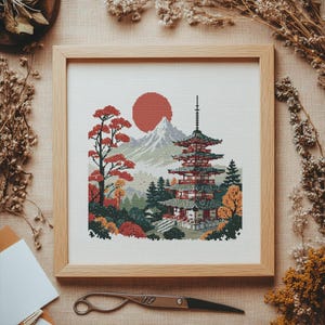 Può includere: Ricamo a punto croce di una pagoda giapponese, sole rosso e paesaggio montano. L'opera è incorniciata in una cornice di legno chiaro. La scena comprende alberi e fogliame nei toni del rosso, verde e arancione. L'opera è su uno sfondo neutro.