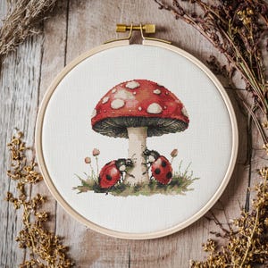Pode incluir: Um aro de bordado de ponto cruz com um cogumelo vermelho e branco com duas joaninhas vermelhas no caule. O cogumelo é cercado por grama verde e pequenas flores brancas.