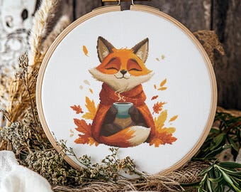 May include: Cross-stitch embroidery in a wooden hoop features a smiling fox wrapped in a cozy orange shawl, holding a blue mug. The fox is surrounded by autumn leaves in shades of orange and yellow. The embroidery is set against a rustic wooden background.